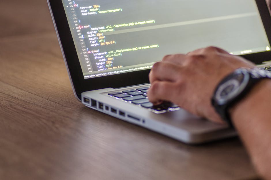 The image shows a person's hand with a wristwatch, using a laptop on a wooden desk. The laptop screen displays lines of code with various colored syntax highlighting, indicating programming or web dev